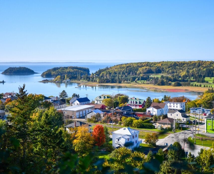 Une petite ville aux maisons blanches et colorées se trouve près d'une baie calme le long de la route touristique des navigateurs à vélo, entourée d'arbres aux feuilles vertes et colorées par l'automne, sous un ciel bleu clair. Des îles et un rivage lointain sont visibles de l'autre côté de l'eau. - Ekilib