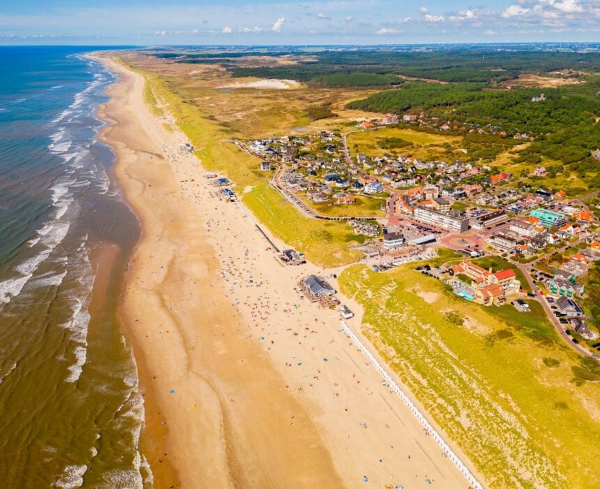 Vue aérienne d'une plage de sable en Hollande du Nord, avec de petites vagues et une ville côtière entourée de végétation verte et de champs de fleurs lointains. Des gens se promènent sur le rivage, certains empruntant des pistes cyclables qui se faufilent dans le paysage et s'étendent au loin. - Ekilib
