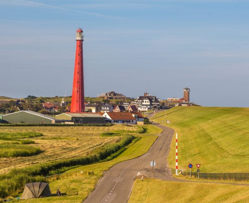 Une route pavée mène à une petite ville avec un grand phare rouge bien en vue, entouré de champs de fleurs et de bâtiments sous un ciel bleu clair. - Ekilib