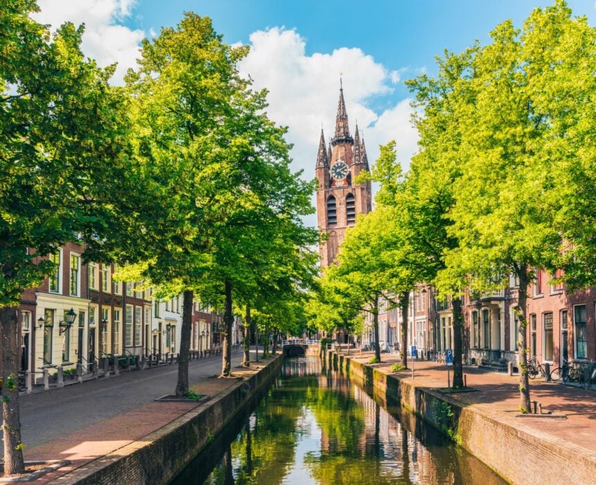 Un canal étroit court entre des rangées de bâtiments et des arbres verts, menant à une grande église historique avec une haute tour d'horloge sous un ciel partiellement nuageux, capturant le charme intemporel des villes historiques comme Amsterdam - où les promenades en vélo sont un plaisir quotidien. - Ekilib