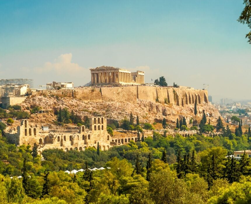 L'image montre l'Acropole d'Athènes, avec le Parthénon au sommet et des ruines anciennes entourées de verdure, sous un ciel bleu clair avec le paysage urbain en arrière-plan. - Ekilib