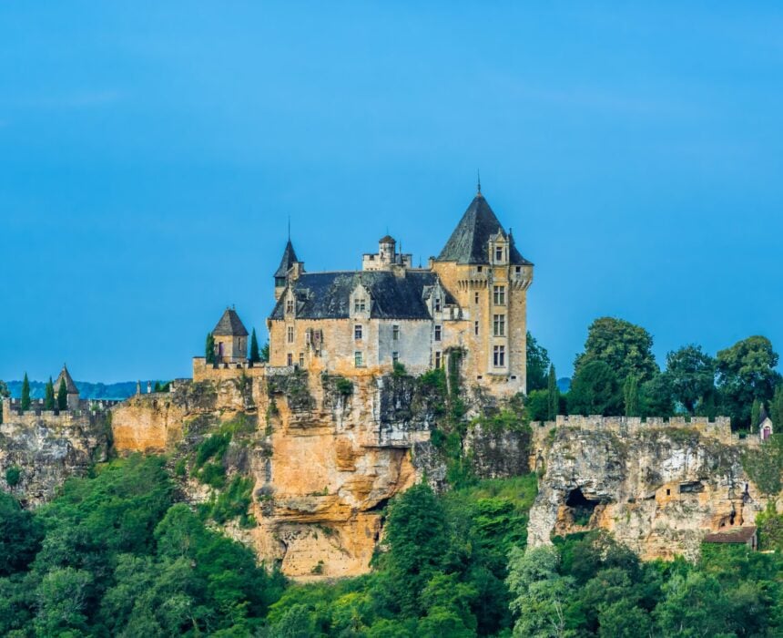 Un grand château de pierre avec de multiples tourelles et des toits abrupts se trouve sur une falaise rocheuse au cœur du Périgord, entouré de verdure sous un ciel bleu clair - un spectacle parfait pour toute aventure à vélo en Dordogne. - Ekilib
