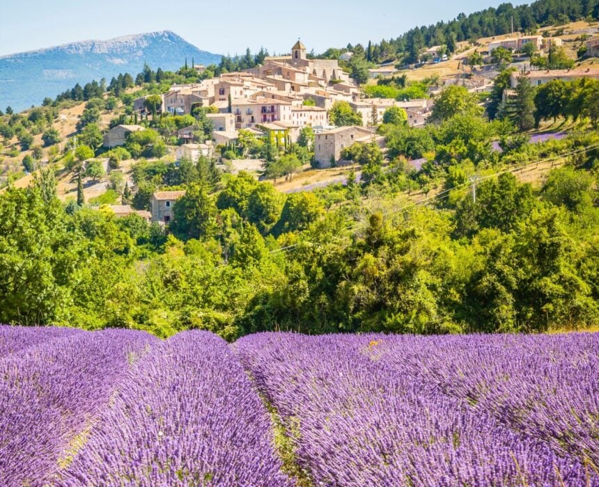 Un champ de lavande en fleurs s'étend au premier plan, tandis qu'un village à flanc de colline et des arbres verts évoquent le charme de la Provence - idéal pour une aventure en vélo-croisière ou en bateau sous un ciel clair et des montagnes lointaines. - Ekilib