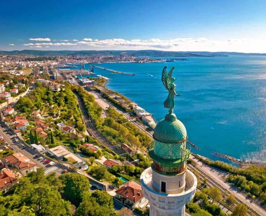 Un grand phare au dôme vert et à la statue ailée se dresse près d'une côte le long de la véloroute Venise-Trieste-Porec, surplombant les eaux bleues, les routes, les voies ferrées et une ville qui s'étend au loin sous un ciel dégagé. - Ekilib