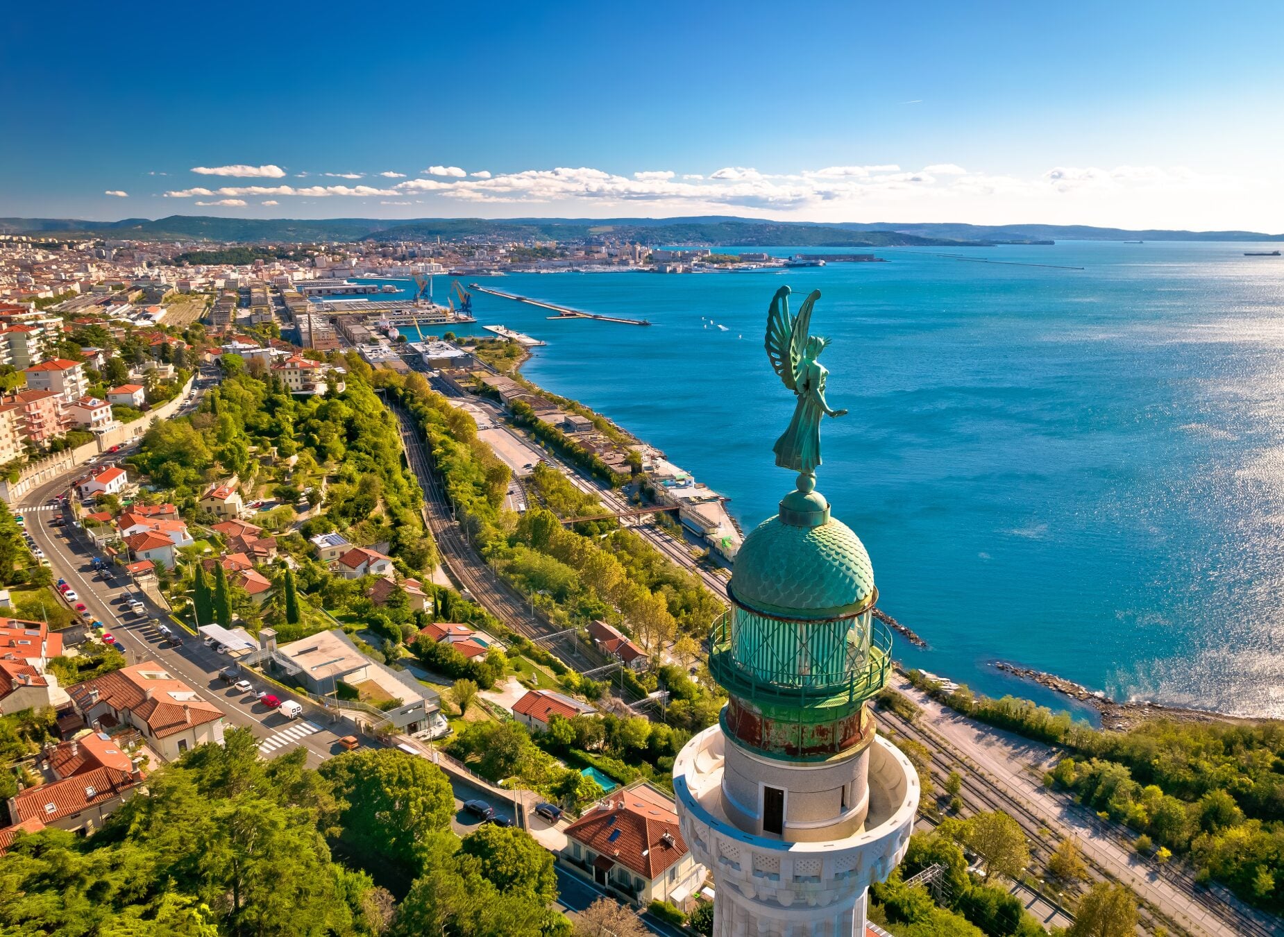 Un grand phare au dôme vert et à la statue ailée se dresse près d'une côte le long de la véloroute Venise-Trieste-Porec, surplombant les eaux bleues, les routes, les voies ferrées et une ville qui s'étend au loin sous un ciel dégagé. - Ekilib