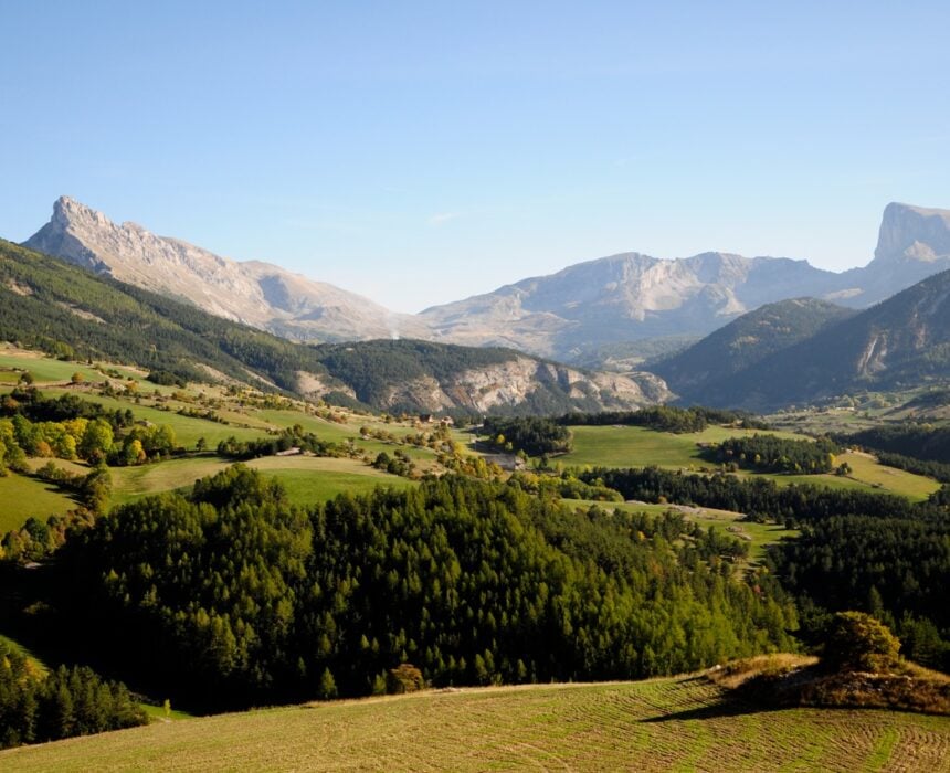 Une large vallée avec des champs verts, des arbres et des collines ondulantes, entourée de hautes montagnes escarpées sous un ciel bleu clair - un paysage idyllique parfait pour une traversée des Alpes à vélo, éclairé par le soleil et s'étendant au loin. - Ekilib