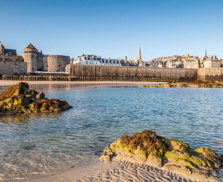 Vue d'une plage de sable avec des rochers dans l'eau peu profonde, face à l'enceinte fortifiée et aux bâtiments historiques de Saint-Malo, en Bretagne, sous un ciel bleu clair, idéal pour une découverte à vélo. - Ekilib