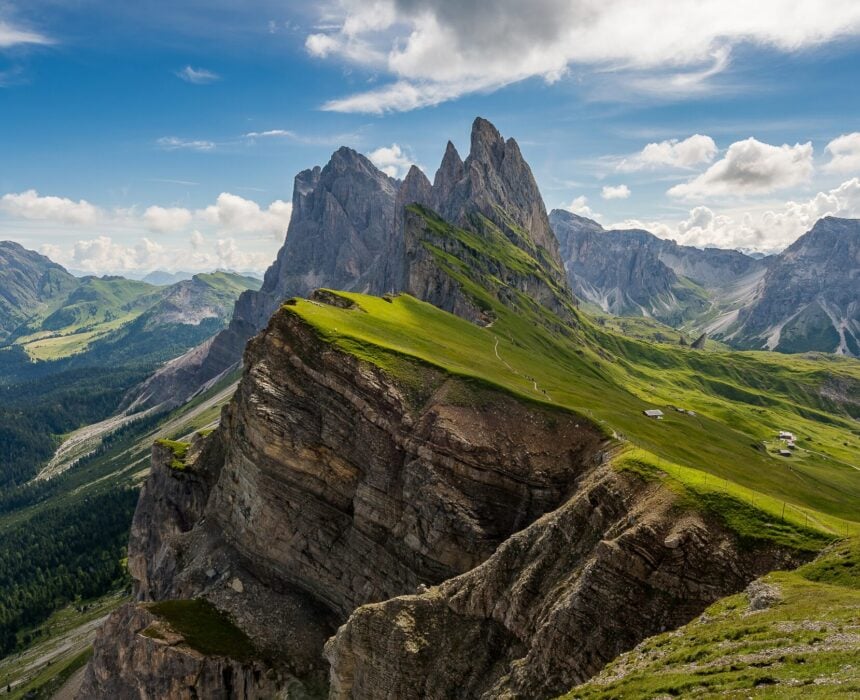 Dans les Dolomites de Velo, des pics montagneux déchiquetés s'élèvent au-dessus de pentes vertes et herbeuses parsemées d'affleurements rocheux. De petites constructions ou des cabanes éparses parsèment le flanc de la colline, sur fond de ciel bleu avec des nuages en arrière-plan. - Ekilib