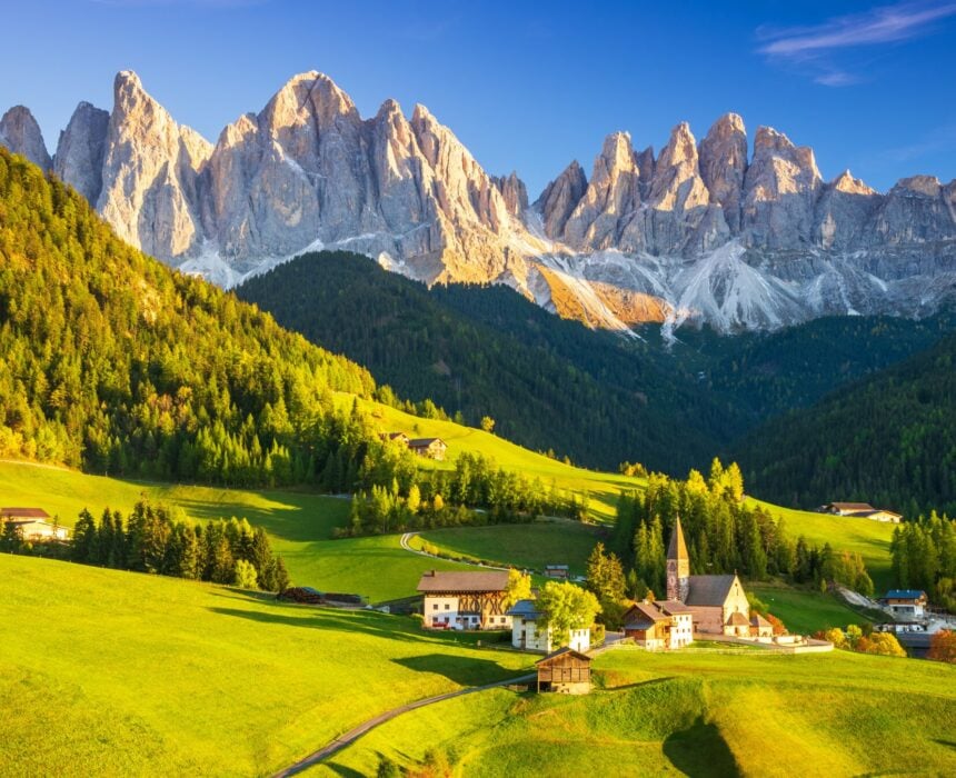 Un petit village aux maisons éparses et à l'église se trouve dans une vallée verdoyante entourée d'arbres dans les Dolomites, avec des pics montagneux déchiquetés s'élevant derrière, invitant les amateurs de vélo sous un ciel bleu clair. - Ekilib