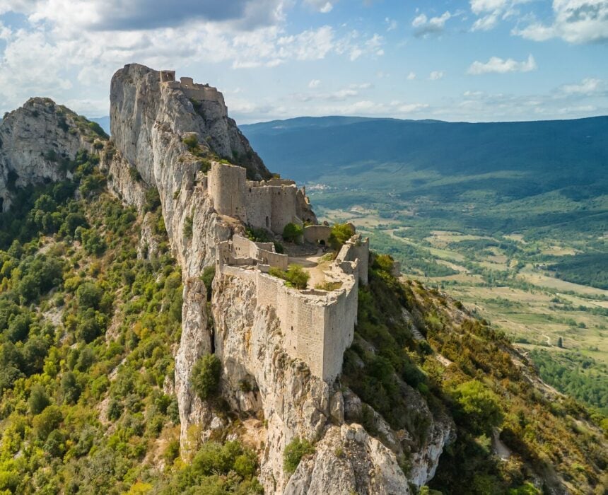 Vue aérienne d'une ancienne forteresse en pierre, aux murs partiellement en ruine, construite sur une crête rocheuse, entourée de collines verdoyantes et d'une large vallée - un paysage parfait pour ceux qui explorent la région en canal du midi vélo sous un ciel partiellement nuageux. - Ekilib