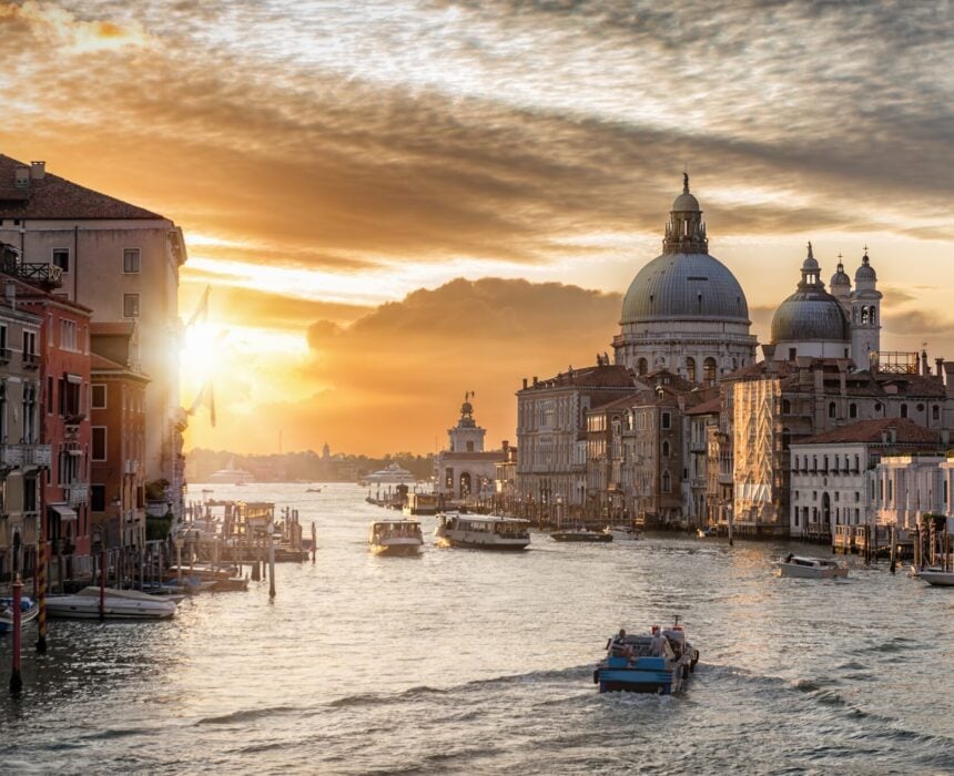 Vue du Grand Canal de Venise au coucher du soleil, avec des vélos et des bateaux glissant sur l'eau, des bâtiments historiques bordant les rives et la basilique Santa Maria della Salute visible sous un ciel partiellement nuageux. - Ekilib