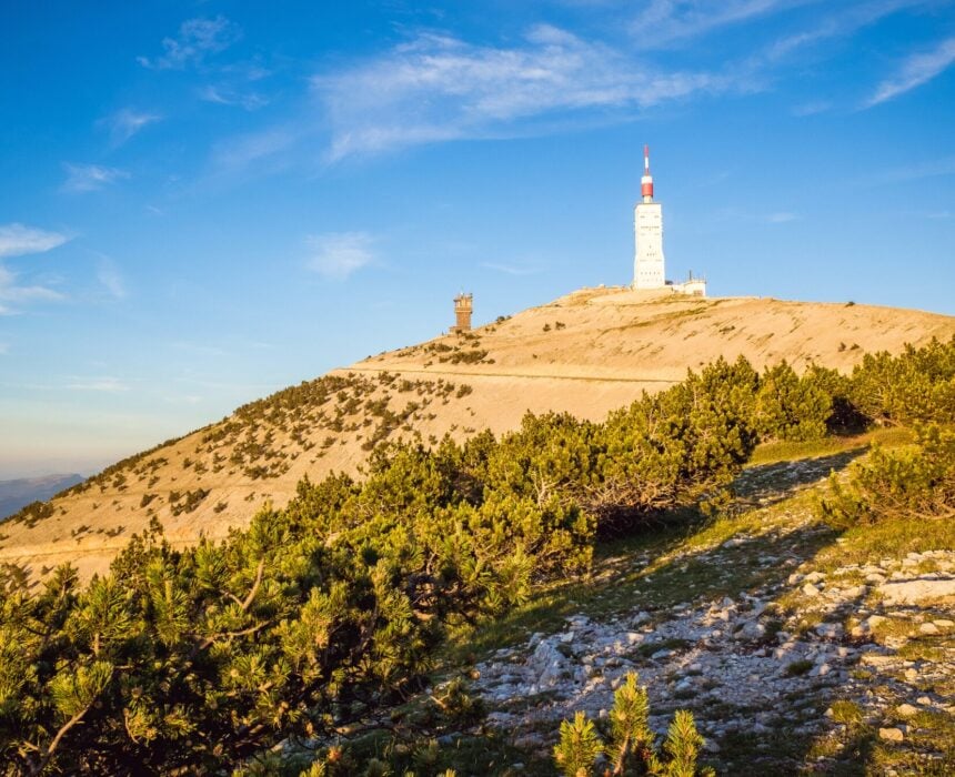Une colline au terrain rocailleux et aux arbustes verts mène à une tour blanche au sommet rouge, qui se détache sur le ciel bleu clair de la Provence et la lumière vive du soleil. Une structure en pierre plus petite se dresse à proximité, reflétant l'esprit du Mont Ventoux, célèbre auprès des amateurs de vélo. - Ekilib