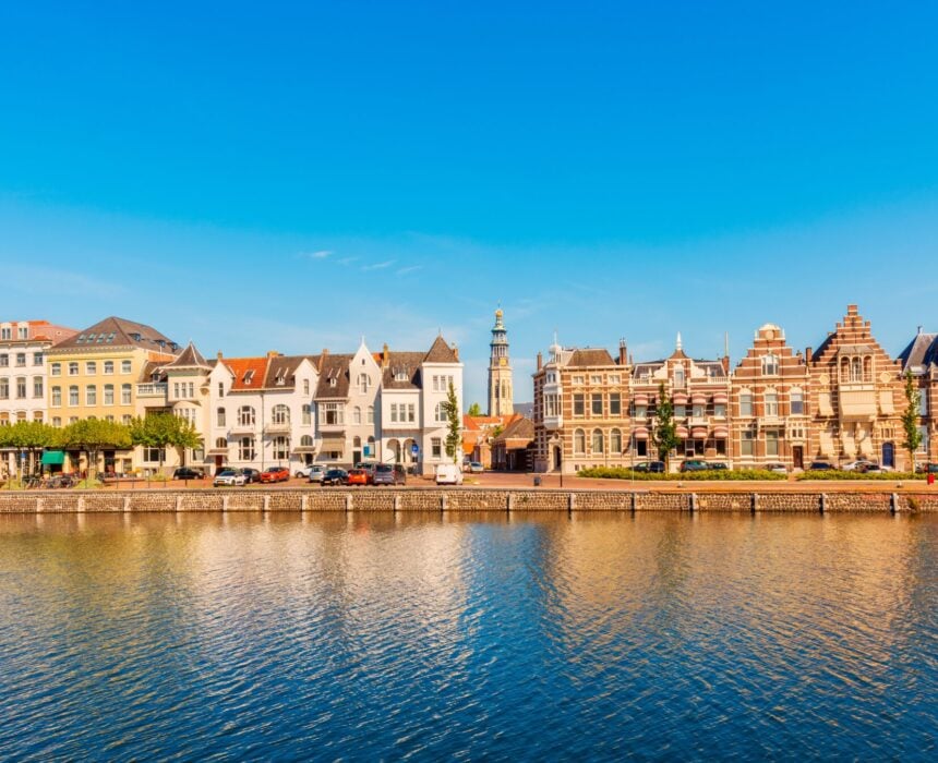 Une rangée de bâtiments historiques aux styles architecturaux variés borde le front de mer d'Amsterdam sous un ciel bleu clair, avec des voitures garées le long de la rue et dont les reflets se reflètent dans l'eau calme. - Ekilib