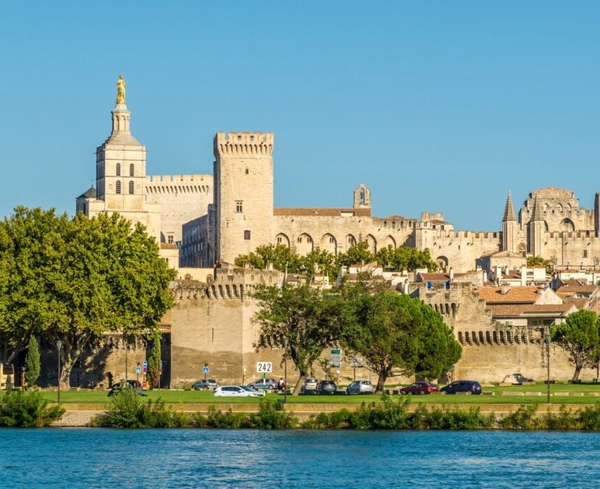 Un grand complexe historique de palais en pierre avec des tours et des tourelles se dresse derrière un mur médiéval, avec au premier plan des arbres, des voitures garées et des cyclistes qui font des tours en vélo en Provence le long de la rivière sous un ciel bleu clair. - Ekilib