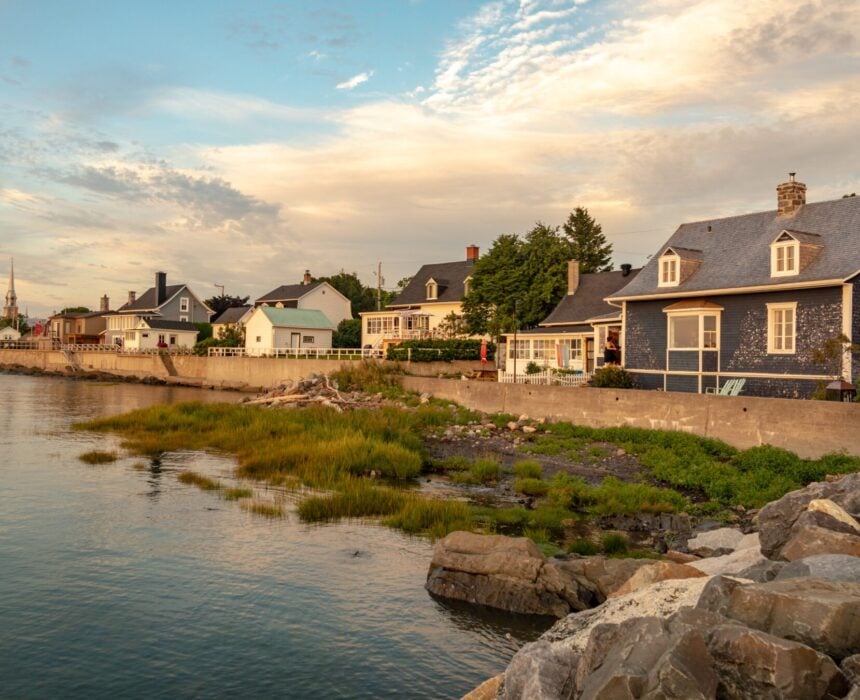 Rangée de maisons au bord de l'eau au coucher du soleil, avec un rivage rocheux et une végétation verte près de l'eau calme ; la scène évoque des souvenirs de la Route des Navigateurs à vélo sous un ciel partiellement nuageux. - Ekilib