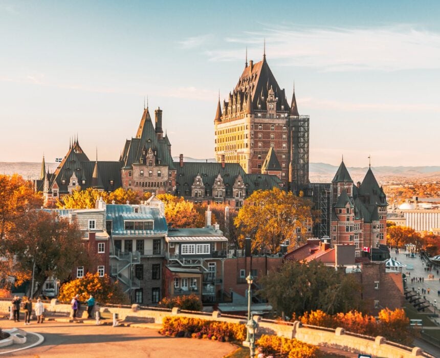 Vue du Château Frontenac à Québec, Canada, près de la route des navigateurs à vélo, entouré de feuillages d'automne et de bâtiments historiques sous un ciel dégagé. On peut voir des personnes marchant au premier plan. - Ekilib
