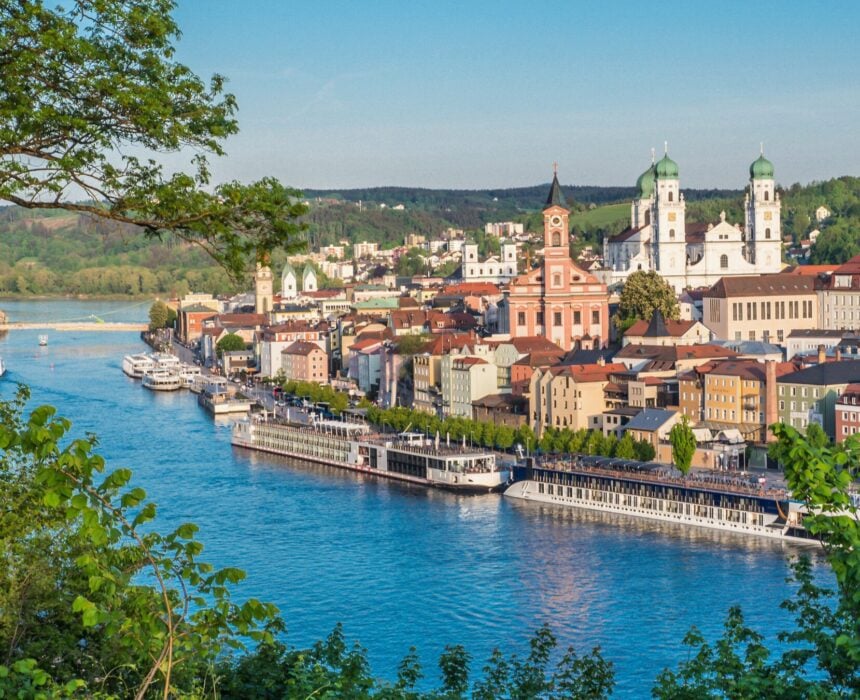 Une ville européenne au bord de la rivière avec des bâtiments colorés, des églises proéminentes aux dômes verts, et des bateaux amarrés le long de la rive - parfait pour un voyage vélo Danube - entourés de verdure et d'arbres sous un ciel bleu limpide. - Ekilib