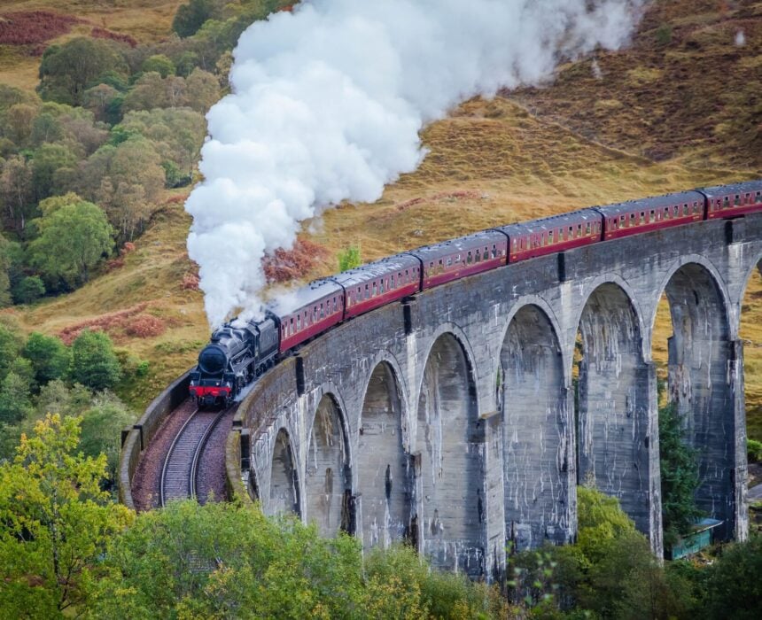 Un train à vapeur noir avec des voitures rouges traverse un grand viaduc en pierre avec de multiples arches, entouré d'arbres verts et de collines herbeuses, en émettant un panache de fumée blanche. - Ekilib