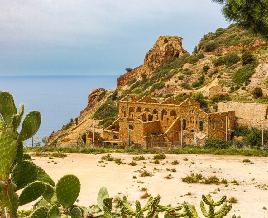 Au pied d'une colline rocheuse proche de la mer, entourée d'une végétation clairsemée et de cactus sous un ciel dégagé, se dresse une bâtisse en pierre délabrée, scène évocatrice d'un voyage en vélo Sardaigne sur la rude Costa Verde. - Ekilib