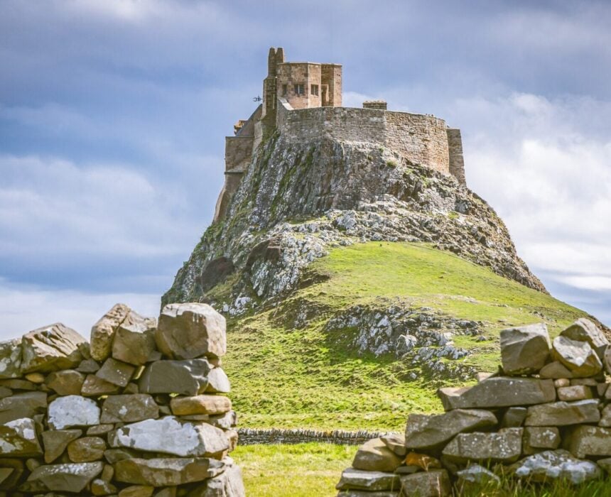 Un château en pierre se trouve au sommet d'une colline rocheuse, entouré d'herbe et d'un ciel partiellement nuageux. Au premier plan, un muret de pierre est percé d'une ouverture. - Ekilib