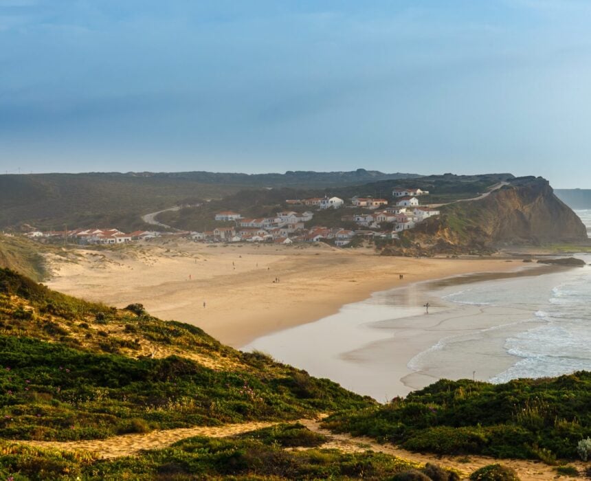 Une plage de sable aux vagues douces borde un village côtier aux maisons blanches et aux toits rouges. Le rivage s'incurve près d'une falaise - un décor parfait pour un tour de l'Algarve en vélo de route - tandis que la verdure remplit le premier plan sous un ciel bleu avec de légers nuages. - Ekilib