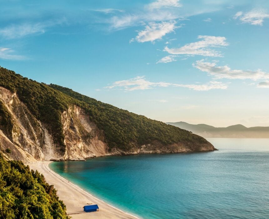 Une large plage de sable aux eaux turquoise s'étire au pied de collines vertes et escarpées, sous un ciel bleu parsemé de nuages. Les cyclistes qui explorent la crète à vélo de route peuvent apercevoir une petite structure bleue près du rivage. - Ekilib
