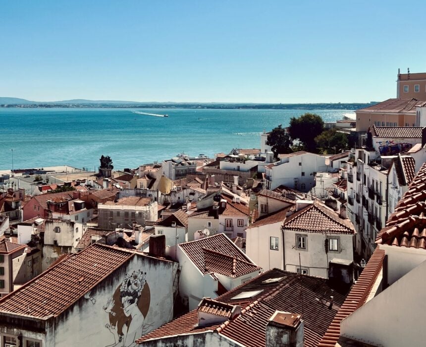 Vue d'une ville côtière avec des toits de tuiles rouges, des bâtiments blancs et une grande peinture murale sur un mur. Un vélo est appuyé contre une clôture, la mer scintille en arrière-plan sous un ciel bleu clair, évoquant le charme de Lisbonne ou de Sagres. - Ekilib