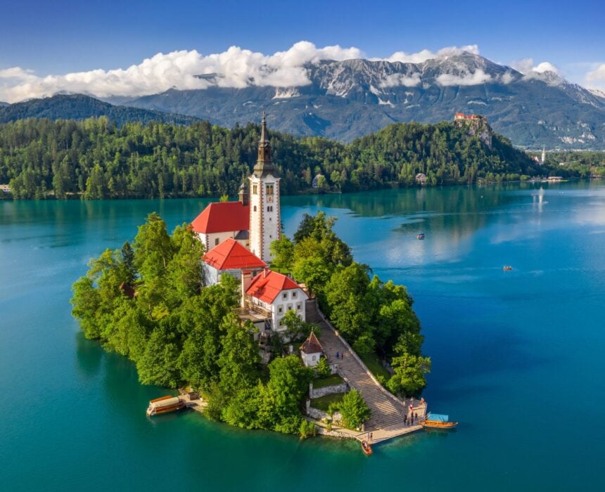 Vue aérienne de l'île de Bled, sur le lac de Bled, en Slovénie, avec l'église de l'Assomption et son haut clocher, entourée d'arbres luxuriants et de collines boisées, parfaite pour les voyageurs qui rêvent d'explorer des endroits comme la vallée du Douro à vélo. - Ekilib