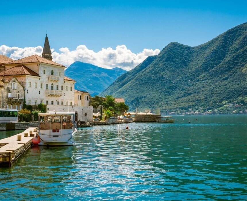 À Monténégro, une ville au bord de l'eau, avec des bâtiments en pierre et des toits de tuiles rouges, se trouve au bord d'une baie calme, parsemée de petits bateaux. Les montagnes vertes s'élèvent à l'arrière-plan sous un ciel bleu clair, idéal pour explorer à vélo et savourer le sens de la liberté des Balkans. - Ekilib