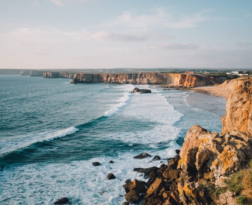 Côte rocheuse avec des vagues s'écrasant sur le rivage, plage de sable en arrière-plan et falaises s'étendant au loin sous un ciel partiellement nuageux - une vue emblématique de la côte atlantique du Portugal à vélo, avec une végétation clairsemée sur les falaises. - Ekilib