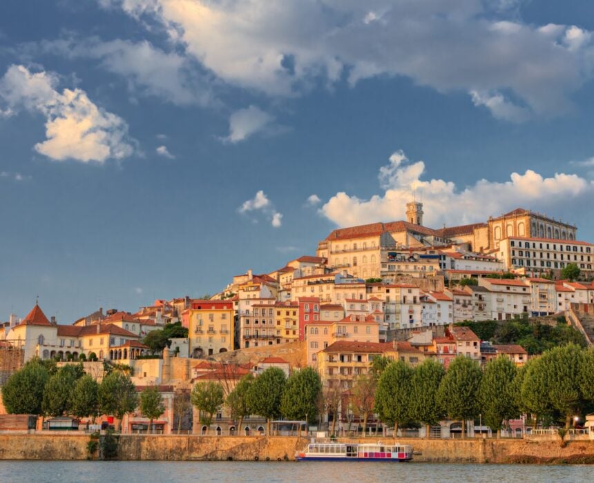 À Porto, un paysage urbain à flanc de colline avec des bâtiments historiques, dont certains avec des toits de tuiles rouges, s'élève au-dessus du fleuve où se trouve un bateau amarré. Des arbres bordent le front de mer sous un ciel partiellement nuageux - un paysage parfait pour une aventure en vélo de route. - Ekilib