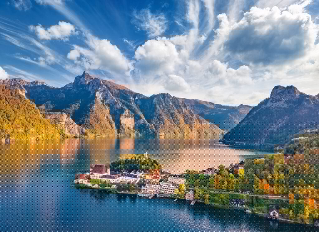 Un petit village se trouve au bord d'un lac le long de la Route des lacs Autriche, entouré d'arbres aux couleurs automnales et de montagnes s'élevant à l'arrière-plan. Le lac reflète le ciel et les sommets, créant un paysage paisible. - Ekilib