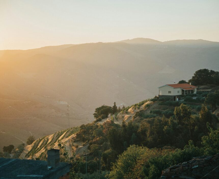 Les maisons sont situées à flanc de colline, entourées d'arbres et de végétation, et surplombent des montagnes et des vallées ondulantes qui rappellent l'exploration à vélo de la vallée du Douro sous un ciel brumeux et doré au coucher du soleil. - Ekilib