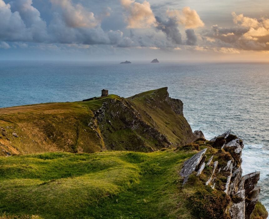Des falaises herbeuses surplombent l'océan sous un ciel partiellement nuageux au coucher du soleil. Lors d'un voyage à vélo en Irlande, découvrez une petite structure en pierre au bord de la falaise, avec deux îles rocheuses visibles dans la mer au loin. - Ekilib