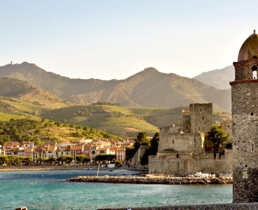 Une ville côtière avec des maisons colorées, un front de mer bordé d'arbres, une tour en pierre avec un dôme, et des ruines historiques en pierre vous attend lors de votre voyage vélo France - avec des collines et des montagnes luxuriantes sous un ciel clair. - Ekilib