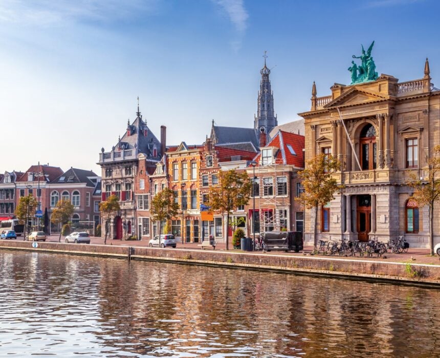 Des bâtiments historiques colorés et une grande structure classique surmontée d'une statue bordent un canal d'Amsterdam sous un ciel bleu, avec des arbres, des vélos garés et le charme des champs de tulipes de Keukenhof à proximité. - Ekilib