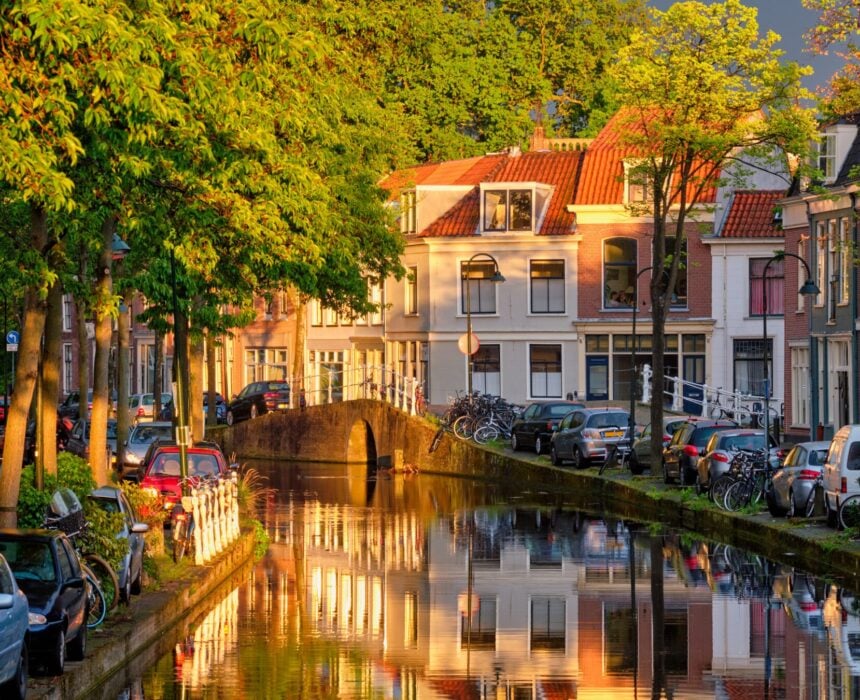 Dans le sud d'Amsterdam, un canal étroit serpente à travers un quartier résidentiel, bordé de voitures garées, de vélos et d'arbres. Des maisons traditionnelles aux toits rouges se reflètent dans l'eau calme où un petit bateau vélo dérive sous la lumière dorée du soleil. - Ekilib