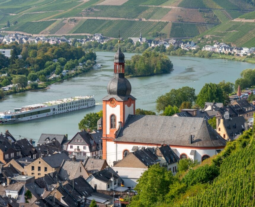 Un bateau de croisière fluviale passe devant une église historique à haut dôme dans une petite ville le long du Rhin, entourée de vignobles et de collines, tandis que d'autres bâtiments urbains et de la verdure bordent les rives de cette croisière pittoresque. - Ekilib