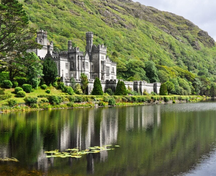 Un grand château de pierre grise se dresse au bord d'un lac calme dans le Connemara, entouré de collines verdoyantes et d'arbres touffus. Son reflet est visible dans l'eau sous un ciel partiellement nuageux - une vue parfaite pour ceux qui partent à la découverte en vélo. - Ekilib