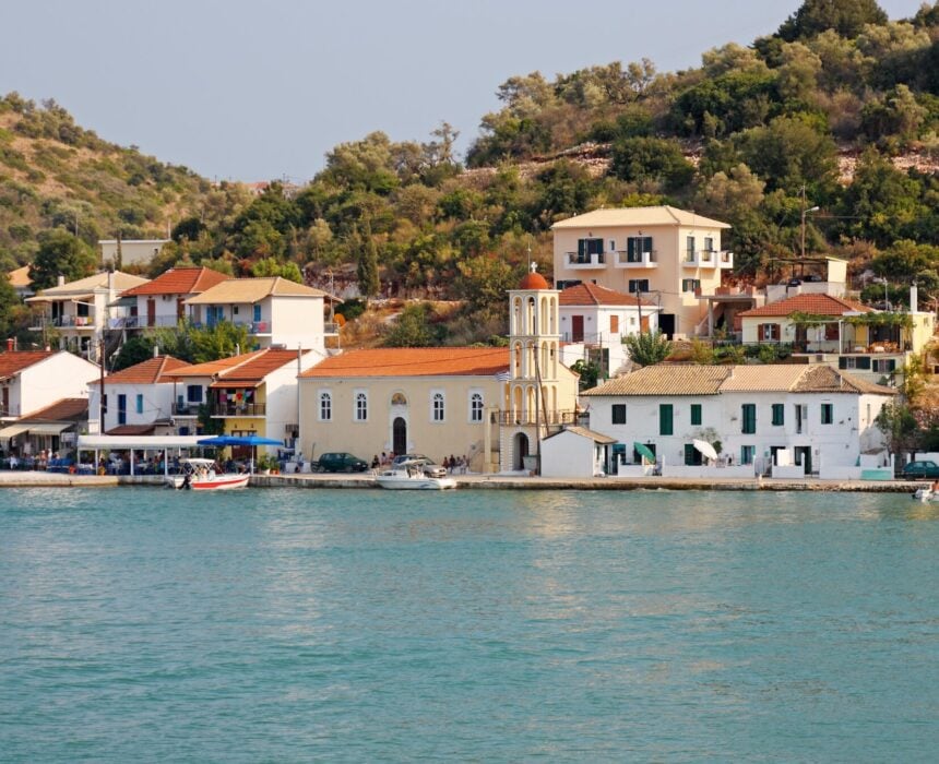 Un petit village côtier aux bâtiments blancs et jaunes, dont une église à clocher, se trouve au bord d'une eau bleue et calme, au pied d'une colline verdoyante couverte d'arbres. Quelques bateaux sont amarrés le long du rivage. - Ekilib