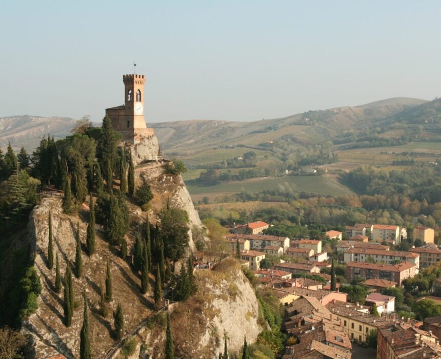 Une tour d'horloge en pierre se dresse au sommet d'une colline rocheuse entourée de cyprès, surplombant une ville aux bâtiments à toits rouges - une scène pittoresque pour ceux qui voyagent de Venise à Florence à vélo à travers des collines vertes et ondoyantes. - Ekilib