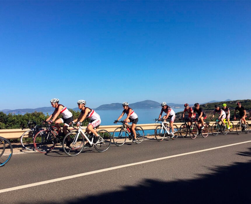 Un groupe de cyclistes roule sur une route pavée le long d'un plan d'eau avec des montagnes au loin sous un ciel bleu clair, profitant de l'aventure de la sardaigne à vélo. Des buissons bordent la route et les cyclistes portent des casques et des vêtements de sport. - Ekilib