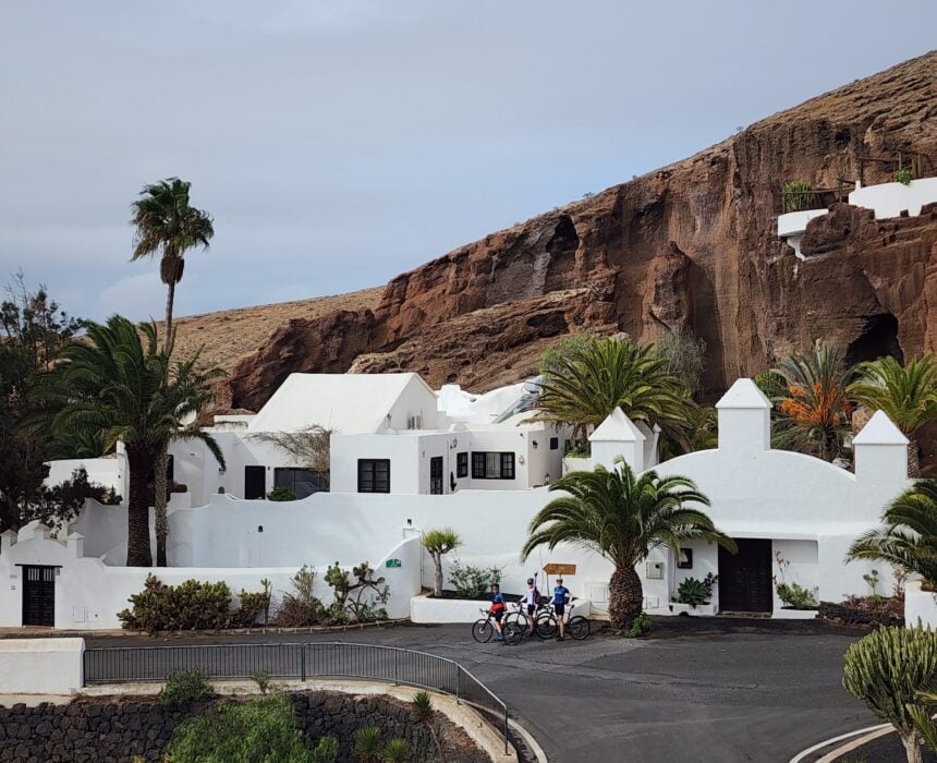 Des bâtiments en stuc blanc avec des entrées voûtées et des palmiers se nichent sur une colline rocheuse à Lanzarote. Trois personnes avec des vélos roulent sur la route incurvée en face, encadrées par un ciel nuageux. - Ekilib