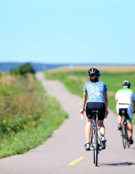 Deux cyclistes casqués empruntent la piste cyclable du Lac Saint Jean dans un paysage rural et verdoyant par une journée ensoleillée. Ils se dirigent vers le lointain, l'herbe et les fleurs sauvages bordant la piste pavée panoramique. - Ekilib