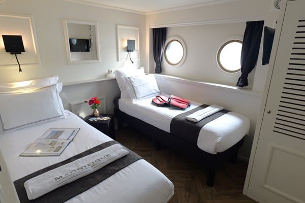 Une petite cabine bien rangée comprend deux lits simples recouverts de draps blancs et de couvertures foncées, une table de chevet ornée de fleurs et de magazines tels que « Hollande Velo », ainsi que deux hublots ronds munis de rideaux sur le mur du fond. Des serviettes sont disposées sur chaque lit. - Ekilib