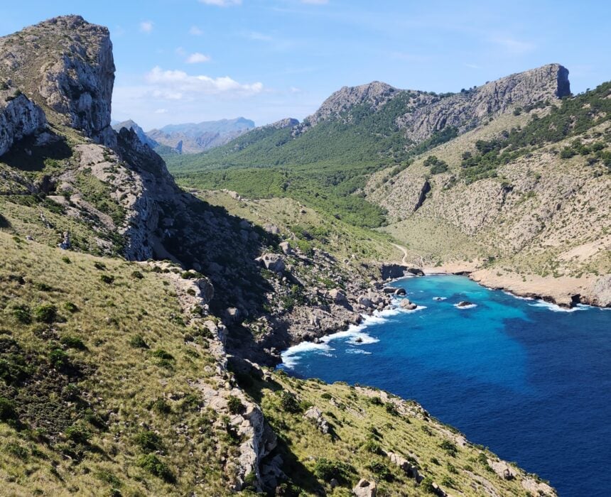 Le long de la route du Majorque, le littoral accidenté présente des falaises rocheuses abruptes, des collines verdoyantes et une petite crique aux eaux bleu foncé et turquoise, sous un ciel partiellement nuageux. Le paysage est essentiellement naturel, sans bâtiments ni personnes visibles. - Ekilib
