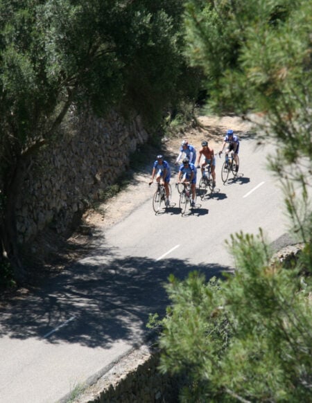 Un groupe de cyclistes d'une agence voyage vélo roule ensemble sur une route étroite et sinueuse bordée de murs de pierre et d'arbres, vue du dessus à travers les branches des arbres. - Ekilib