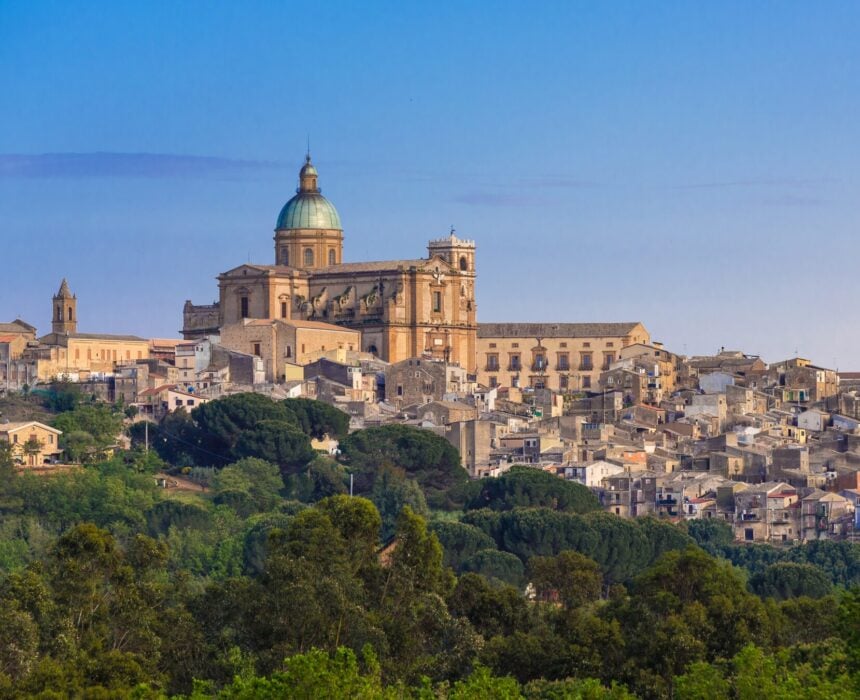 Une grande cathédrale historique au dôme vert surplombe une ville à flanc de colline en Sicile, idéale pour le cyclotourisme, avec des bâtiments en pierre très denses, des arbres verts et un ciel bleu limpide. - Ekilib