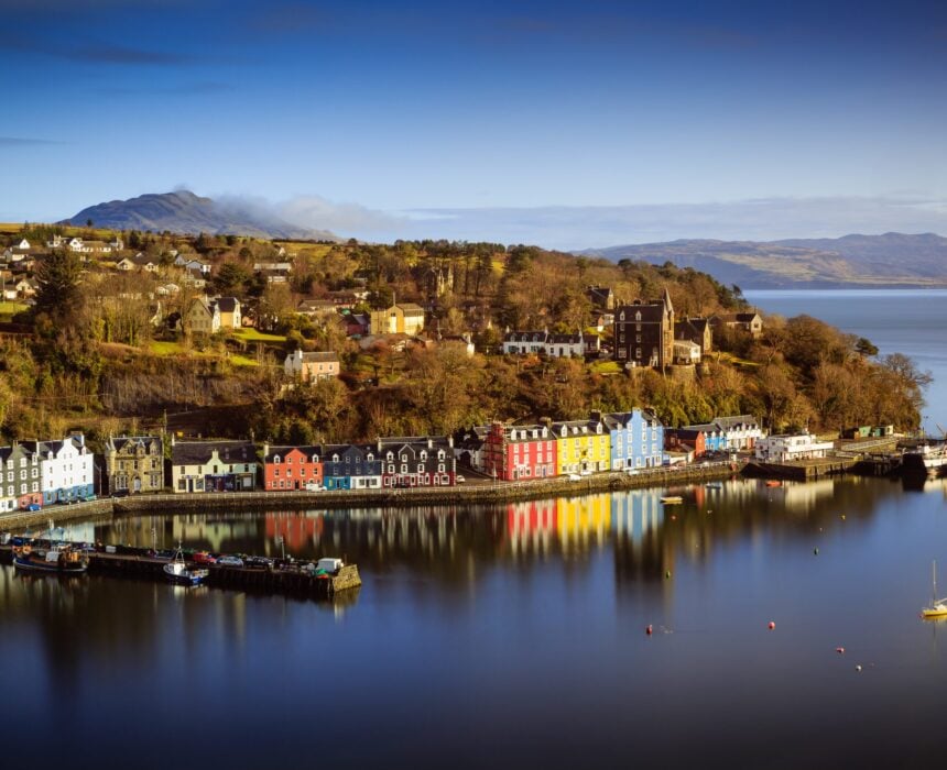 Une rangée de bâtiments colorés borde le front de mer d'une petite ville côtière, où un bateau écossais flotte doucement sur une eau calme, reflétant des scènes vibrantes avec des collines et des maisons éparpillées sous un ciel bleu clair. - Ekilib