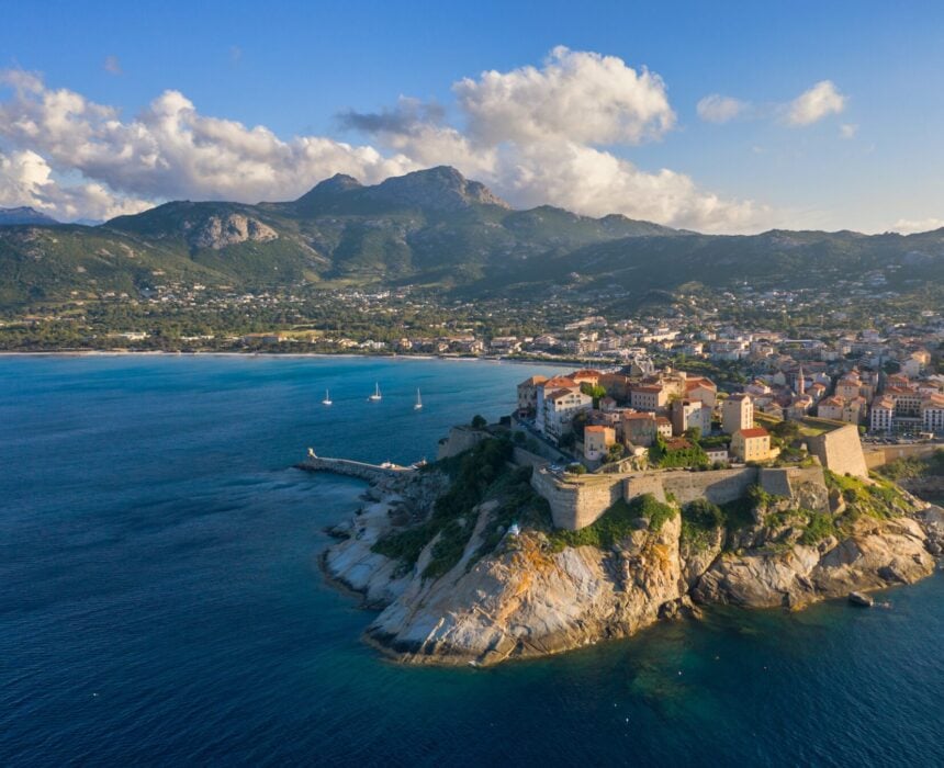 Une ville côtière avec des bâtiments historiques se trouve sur une péninsule rocheuse entourée d'une mer bleue, une étape de la route touristique GT20 vélo Corse. Des montagnes et des paysages verdoyants s'élèvent à l'arrière-plan sous un ciel partiellement nuageux, tandis que des voiliers jettent l'ancre près du rivage. - Ekilib